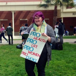 Picture of Amber Melvin with purple hair holding a sign that says 'Pro-Health Pro-Life Pro-Woman Pro-Choice'