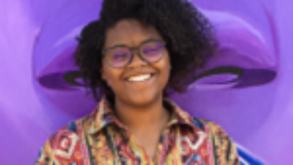 Woman smiles at camera, wearing a colorful blouse, against a purple background.