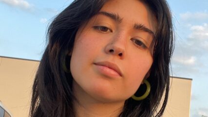 Person with long dark hair looks blankly at camera. Background is a building and a blue sky.