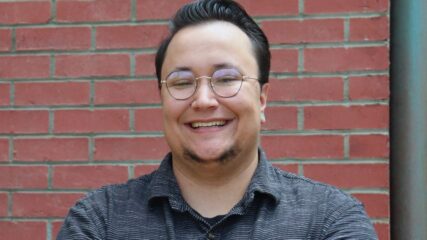 URGE Senior Kansas Organizer, Nigel Morton, stands with crossed arms, smiling, against a brick wall.