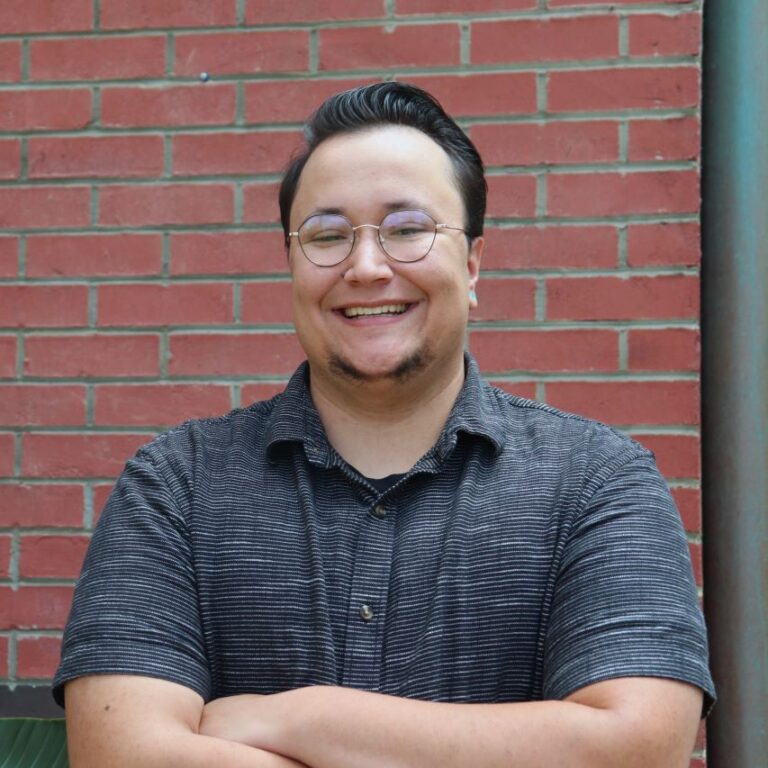 URGE Senior Kansas Organizer, Nigel Morton, stands with crossed arms, smiling, against a brick wall.